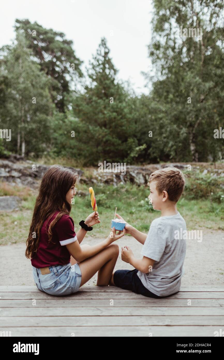 Fratelli mangiando cibo dolce mentre si siedono su pavimento di legno contro albero e cielo Foto Stock