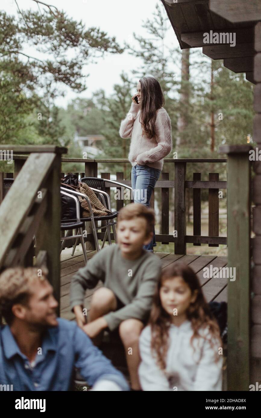 Padre seduto con i bambini sui gradini mentre la donna parla sopra telefono cellulare in portico Foto Stock