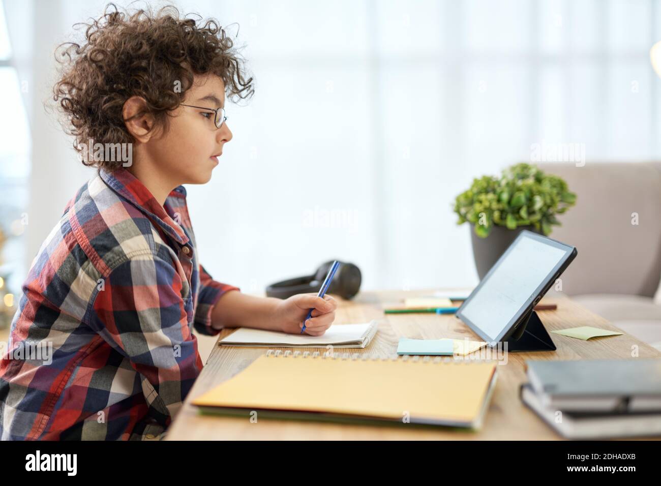 Entusiasta ragazzo della scuola latina che indossa occhiali che fanno appunti, avendo lezioni online utilizzando tablet digitale mentre studia a casa. Formazione online, tecnologia, homeschooling. Vista laterale Foto Stock