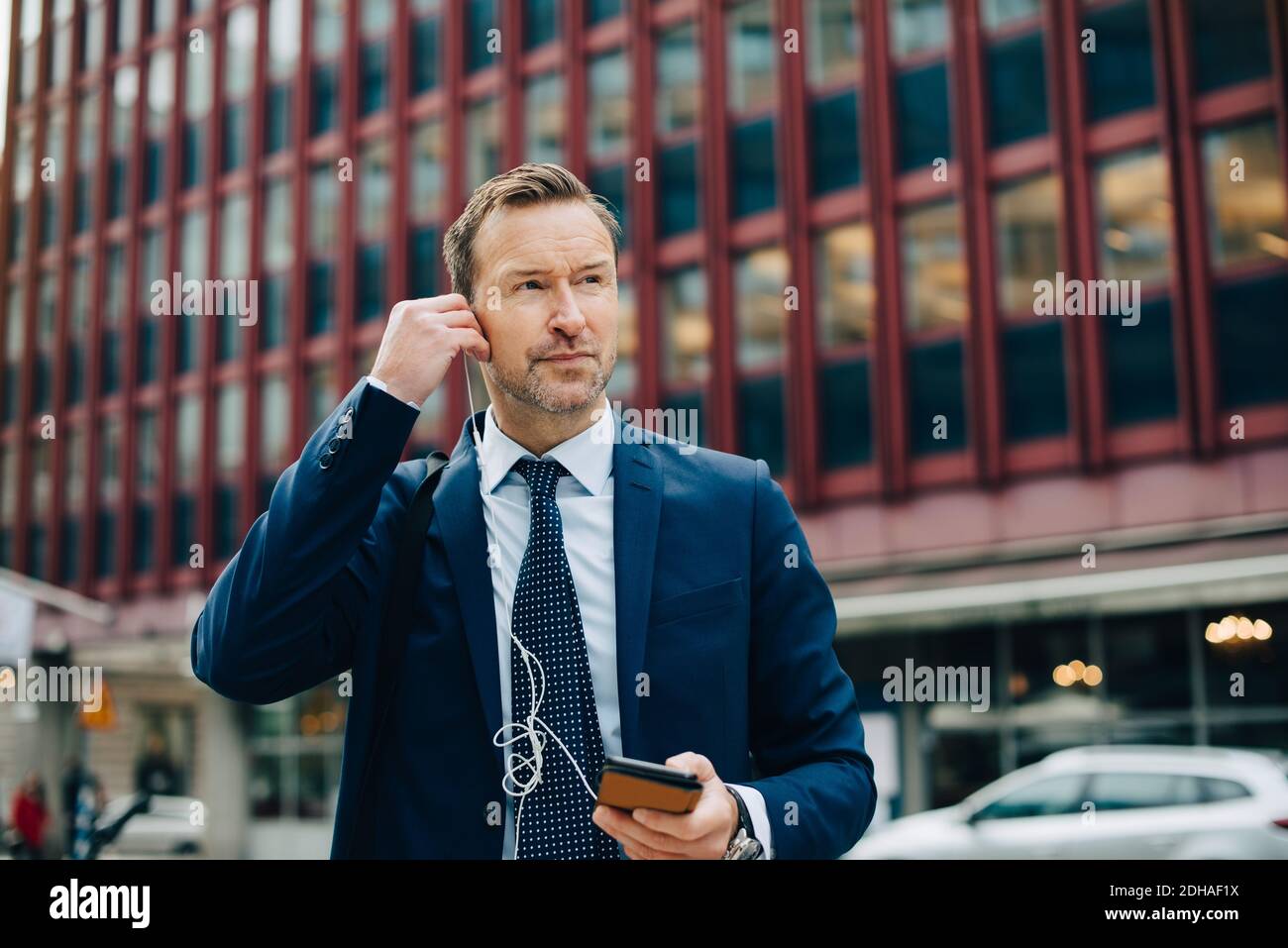 Cuffie intrauricolari per il posizionamento di un uomo d'affari maturo mentre si guarda lontano dagli edifici in città Foto Stock