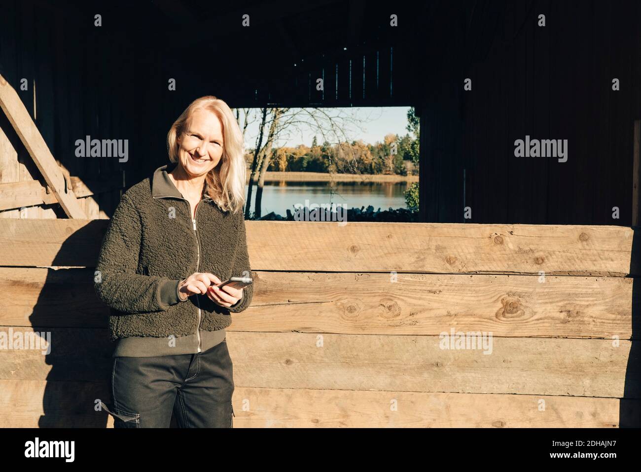 Ritratto di sorridente agricoltore maturo utilizzando il telefono cellulare all'esterno fienile Foto Stock