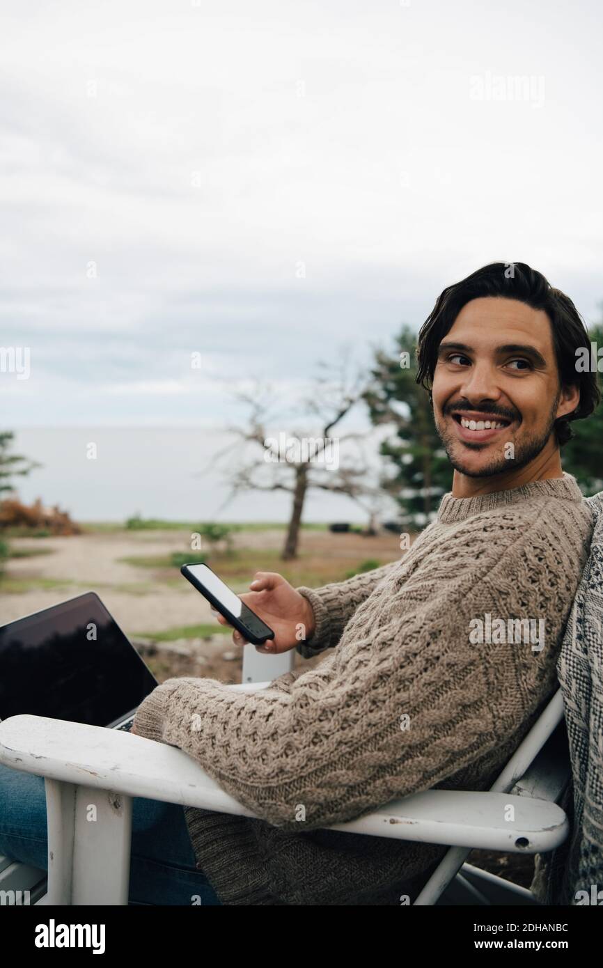 Uomo sorridente con computer portatile e telefono cellulare che guarda lontano seduto sulla sedia contro il cielo Foto Stock