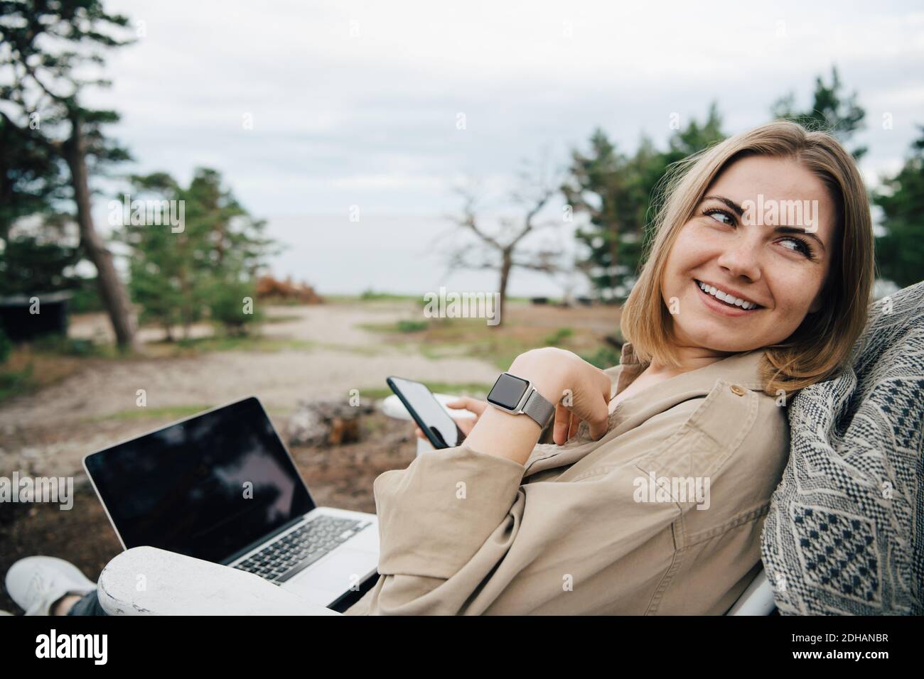 Giovane donna sorridente con tecnologie che guardano lontano mentre si siede sopra sedia durante le vacanze Foto Stock