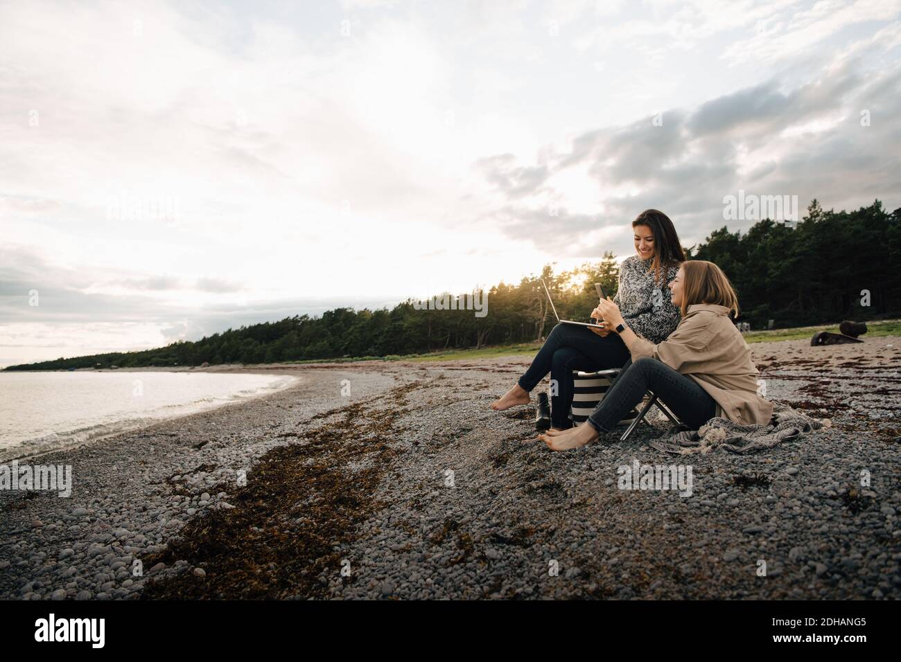 Amici di sesso femminile che guardano il telefono cellulare mentre si siede insieme a. spiaggia al tramonto Foto Stock