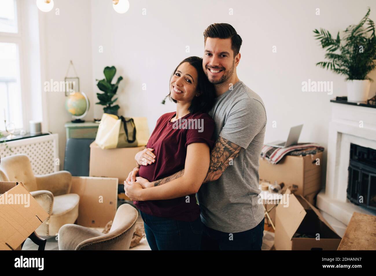 Uomo sorridente che abbraccia una donna incinta mentre si sta in piedi contro le scatole dentro soggiorno Foto Stock