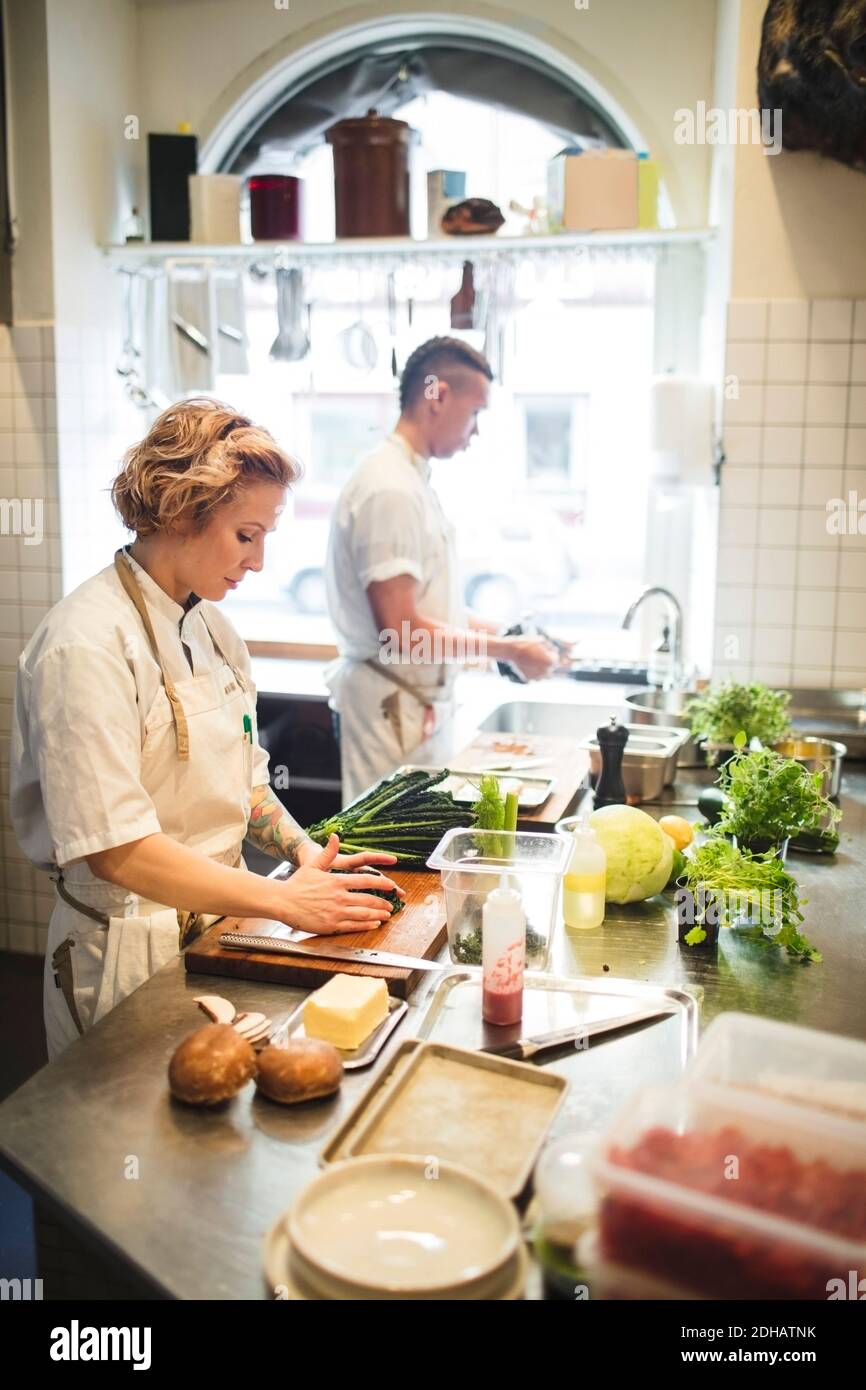 Lo chef femminile sta tagliando le verdure a bordo da un collega che lavora al banco nella cucina del ristorante Foto Stock