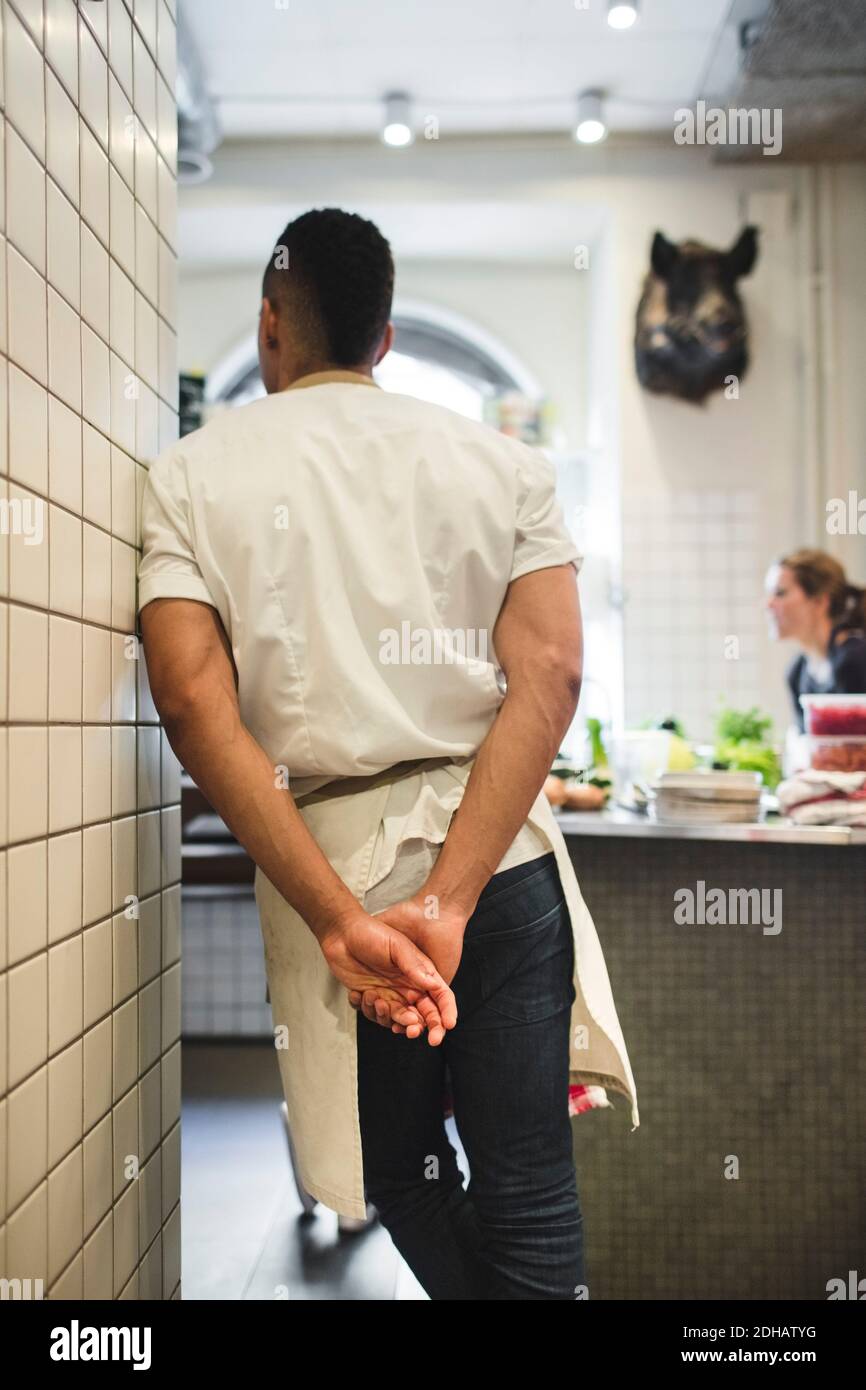 Vista posteriore dello chef maschile in piedi con le mani dietro la schiena a muro in cucina al ristorante Foto Stock