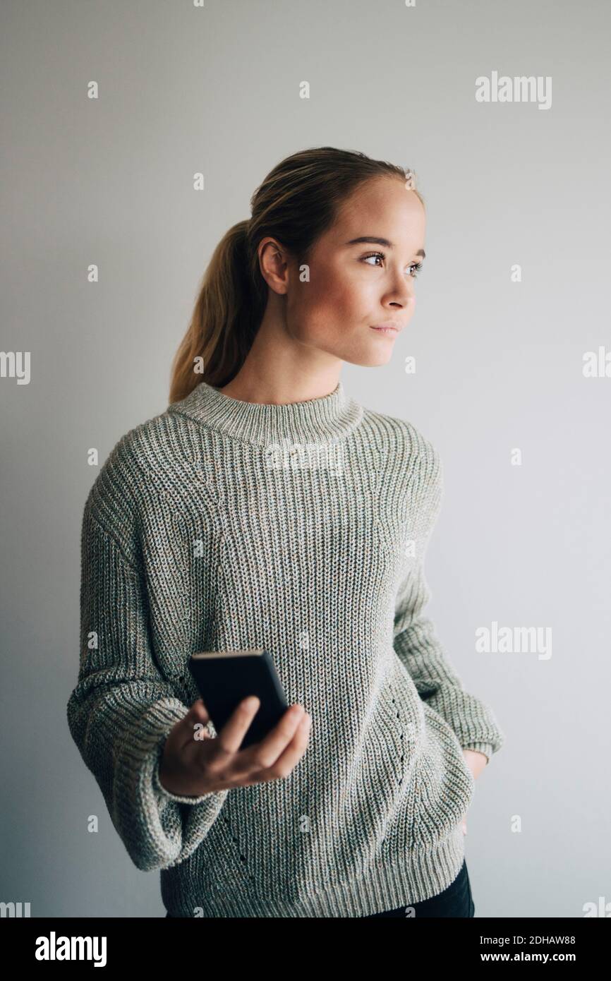 Ragazza adolescente che tiene il telefono cellulare mentre si trova contro la parete bianca a casa Foto Stock