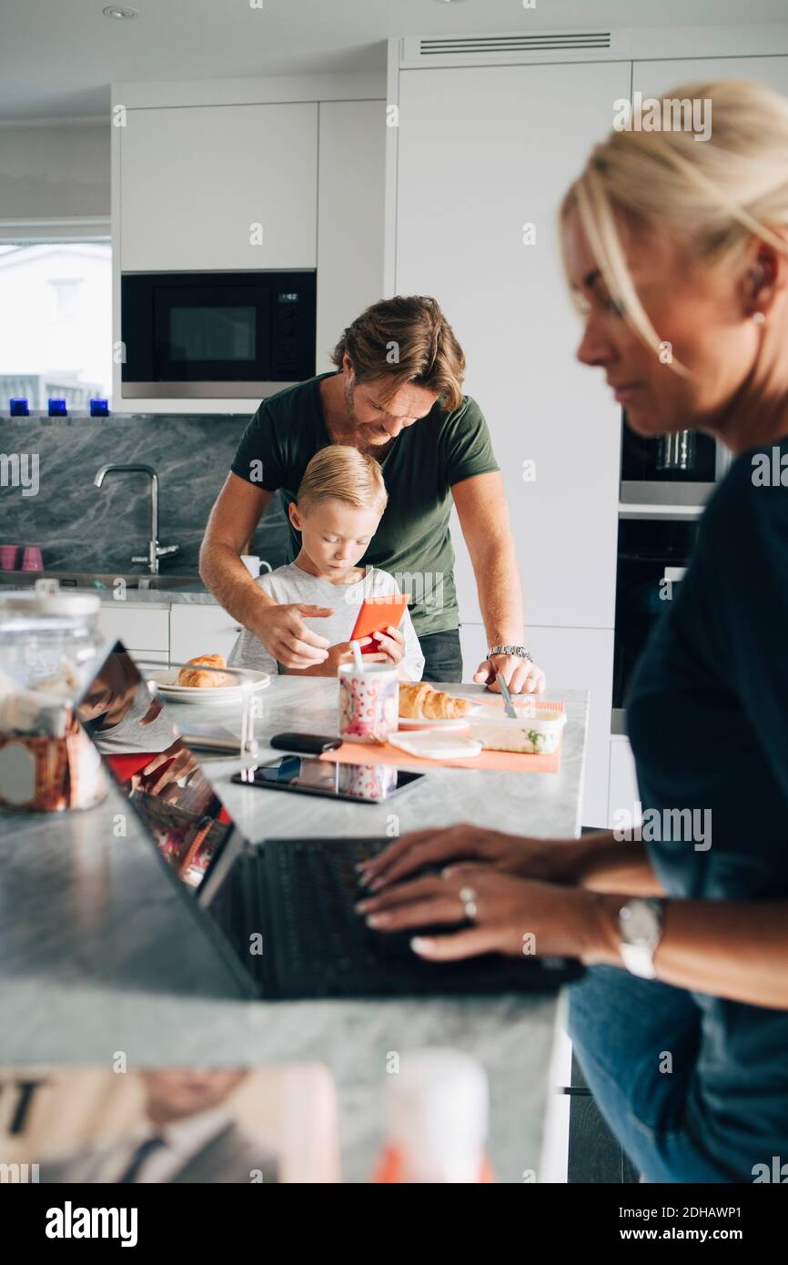 Donna che usa un computer portatile mentre uomo e figlio tengono in mano uno smartphone in cucina a casa Foto Stock