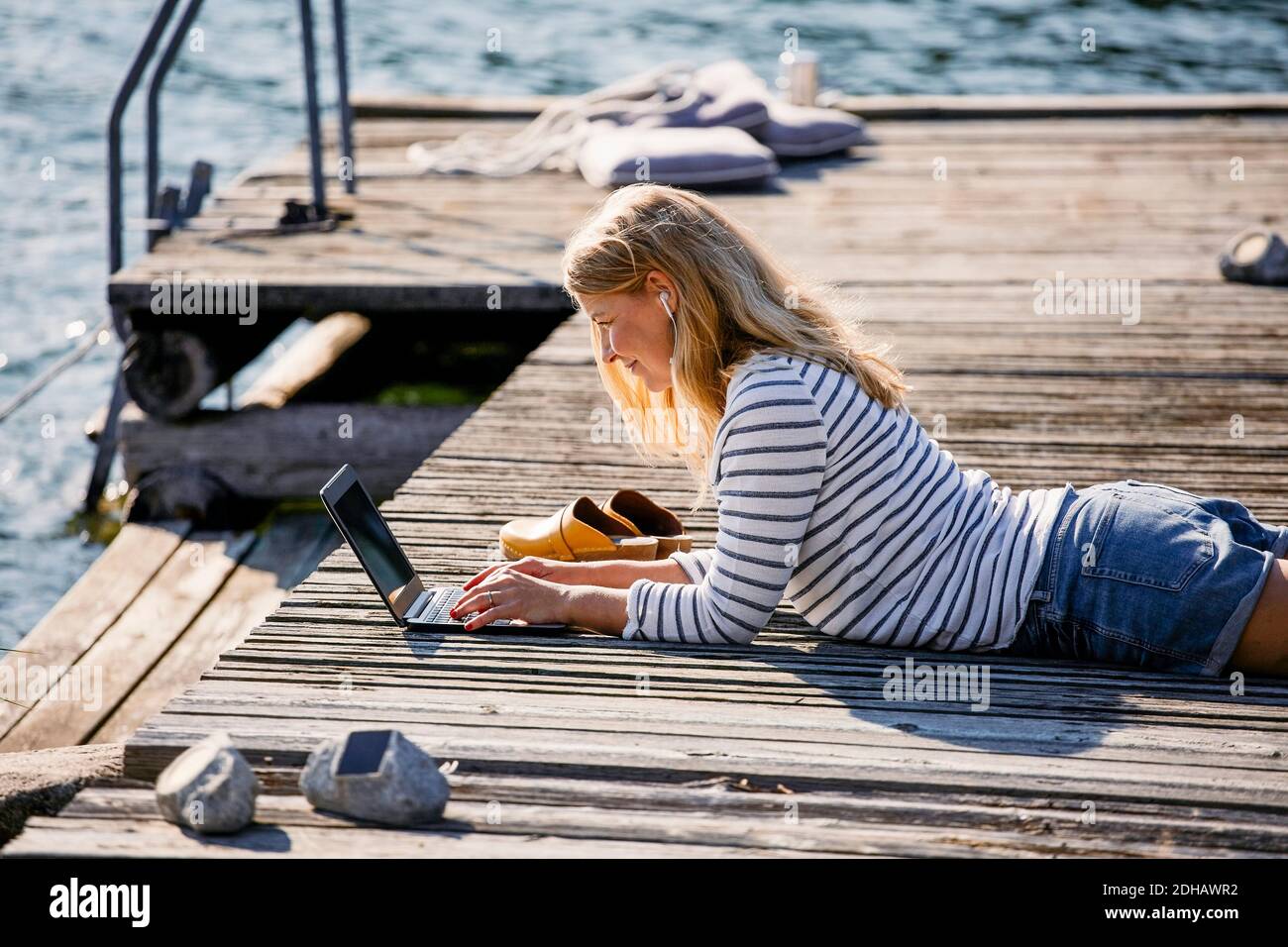 Donna mid adulta che scrive sul computer portatile mentre si trova sopra legno molo durante le vacanze estive Foto Stock