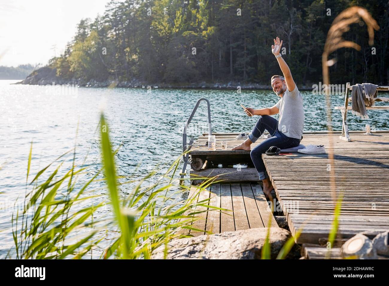 Uomo maturo che guarda via e sventolando mentre tiene il tablet digitale sul molo sopra il lago Foto Stock