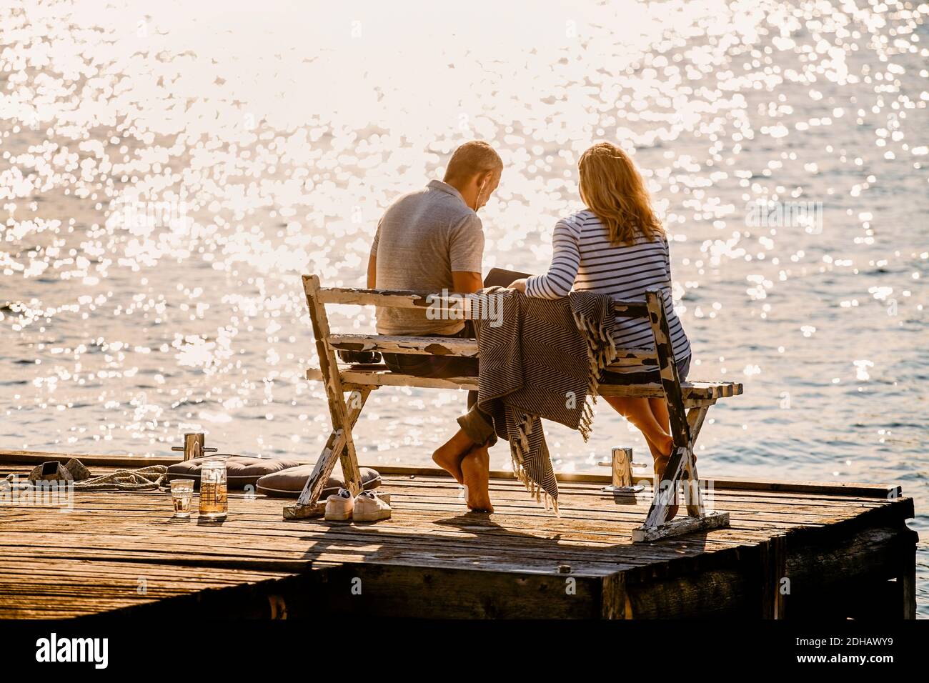 Tutta la coppia che usa il computer portatile mentre si siede sulla panca sopra il molo di legno contro il lago durante le vacanze estive Foto Stock