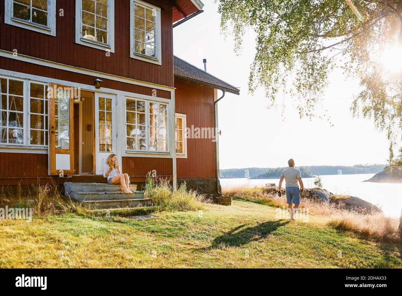 Donna seduta su gradini all'ingresso della cabina di legno mentre uomo che cammina verso il lago il giorno di sole Foto Stock