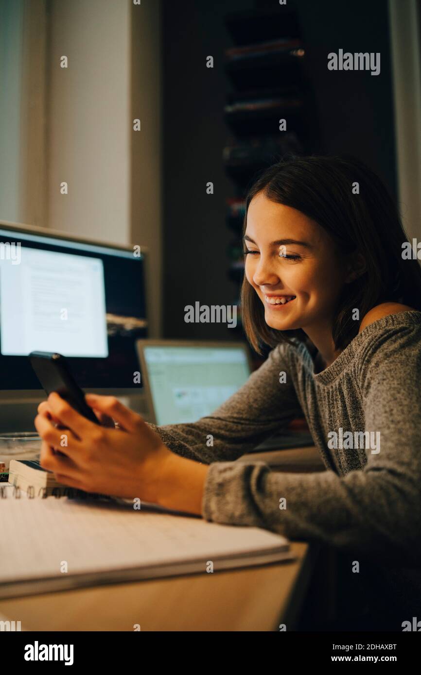 Sorridente ragazza che usa lo smartphone mentre si siede alla scrivania Foto Stock