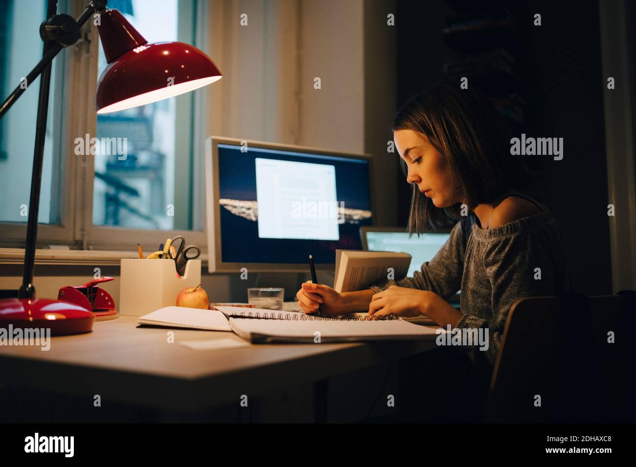 Vista laterale della ragazza che scrive mentre usando il calcolatore a illuminato scrivania Foto Stock
