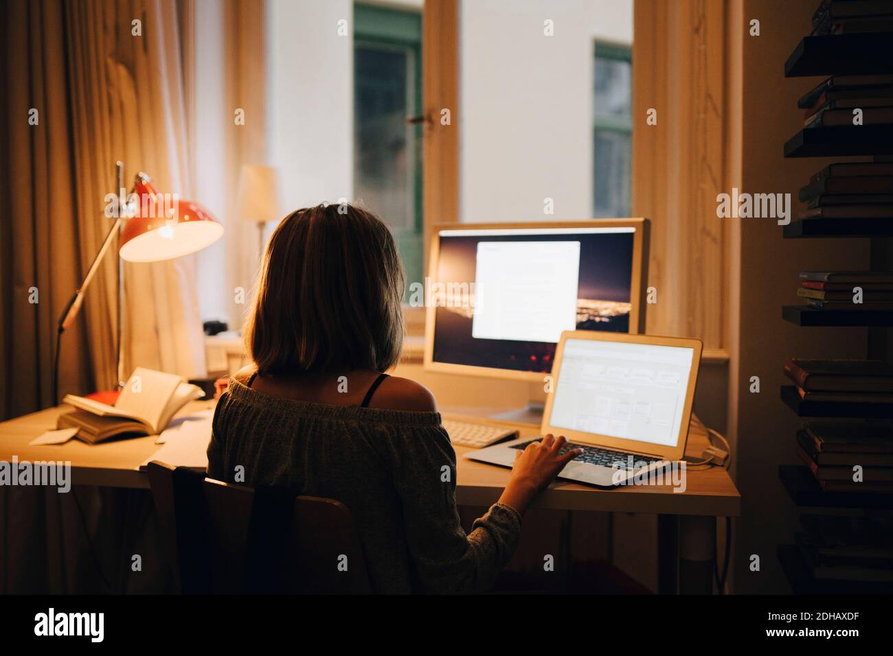 Vista posteriore di una ragazza che utilizza il computer e il computer mentre si è seduti alla scrivania illuminata Foto Stock