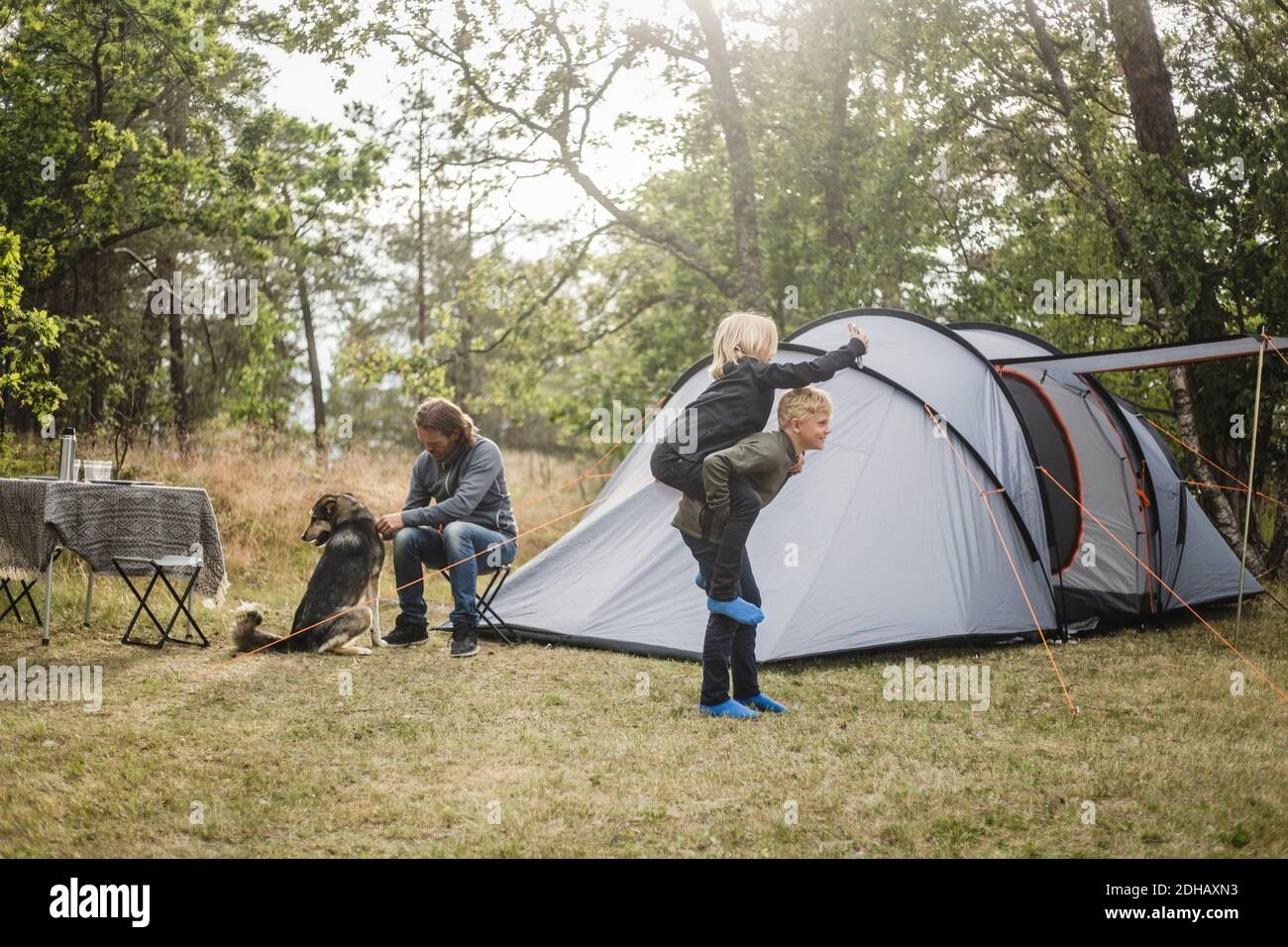 Fratelli che giocano mentre l'uomo seduto con il cane da tenda a. campeggio Foto Stock