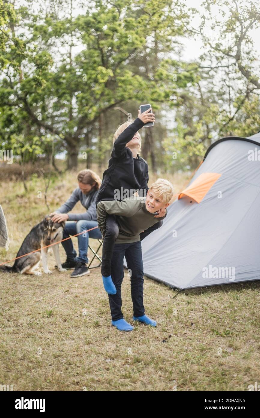 Ragazzo piggybacking sorella con smartphone mentre l'uomo gioca con cane in tenda campeggio Foto Stock