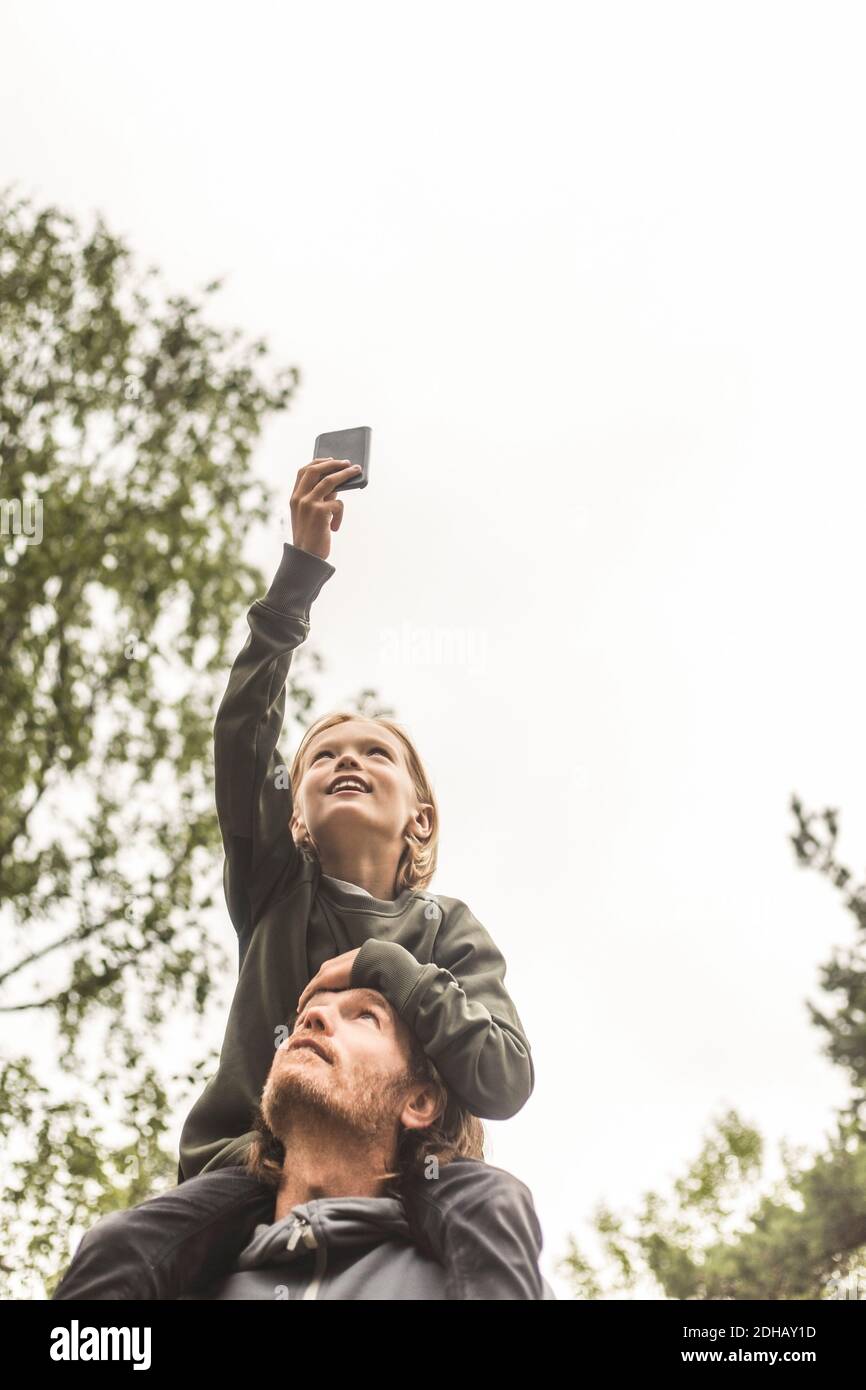 Vista ad angolo basso della ragazza che fotografa attraverso il telefono cellulare mentre seduto sulle spalle del padre contro il cielo Foto Stock