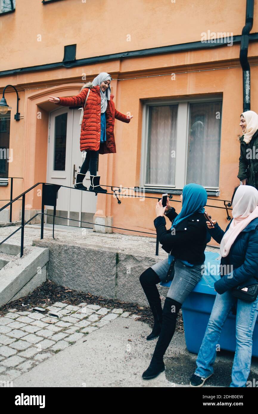Donna con amici che fotografano l'equilibrio femminile sulla ringhiera contro l'edificio in città Foto Stock