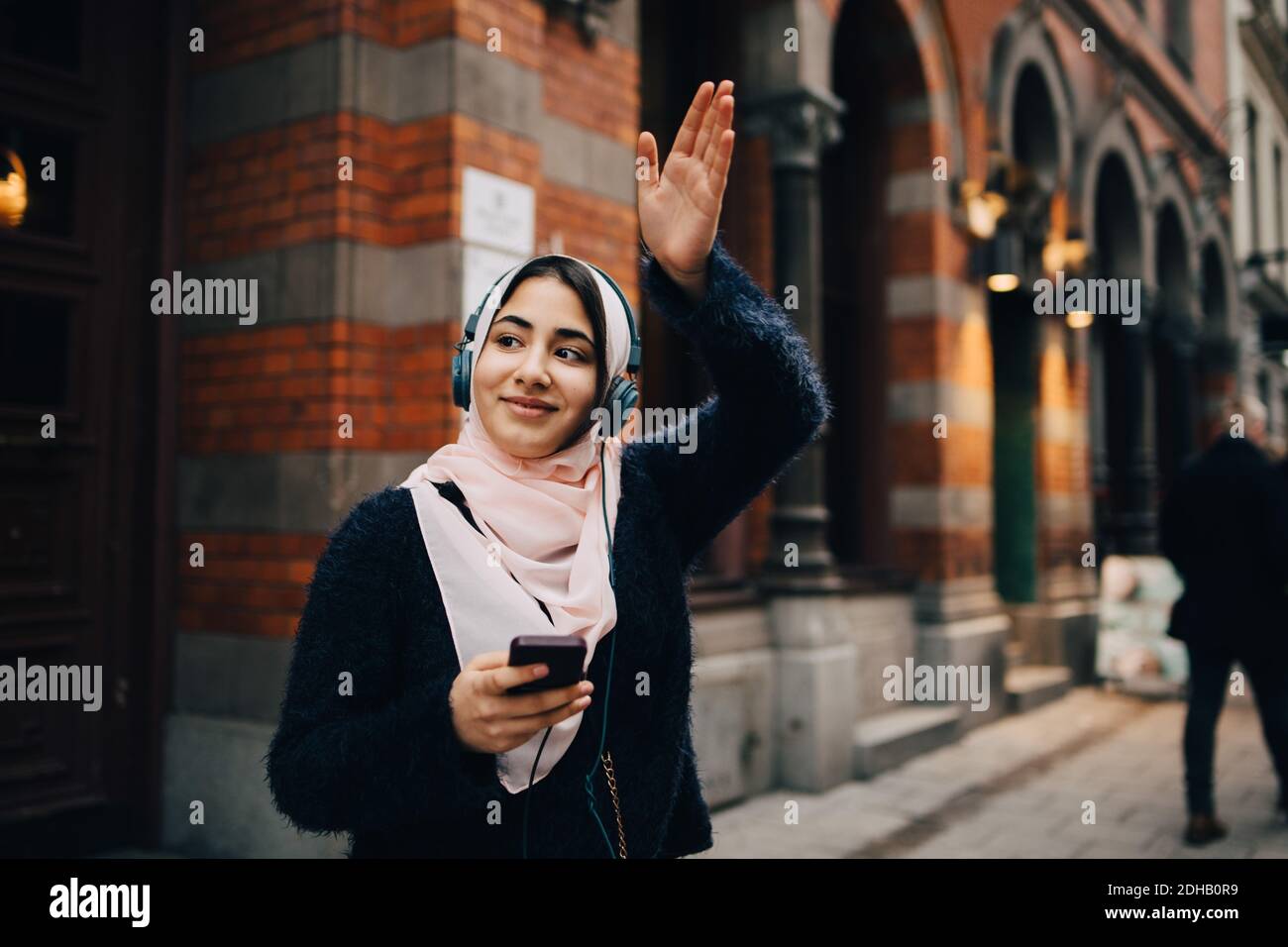 Sorridente ragazza adolescente che agita la mano mentre si ascoltano le cuffie sentiero in città Foto Stock