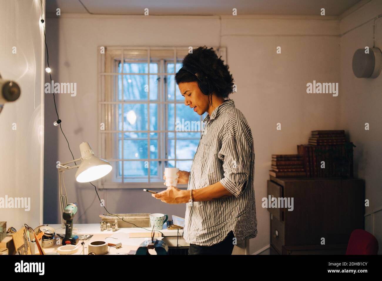 Tecnico con la tazza utilizzando il telefono cellulare mentre si è in piedi alla scrivania in officina Foto Stock