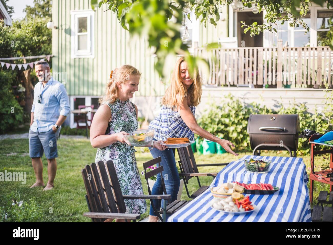 Amici femminili che organizzano il cibo sul tavolo per la riunione sociale dentro cortile durante il fine settimana estivo Foto Stock