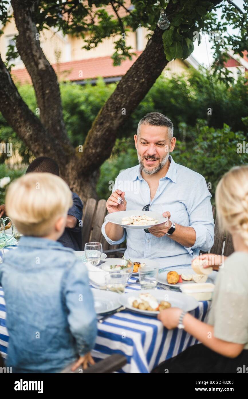 Uomo maturo che parla mentre mostra piatto di cibo ai bambini dentro festa Foto Stock