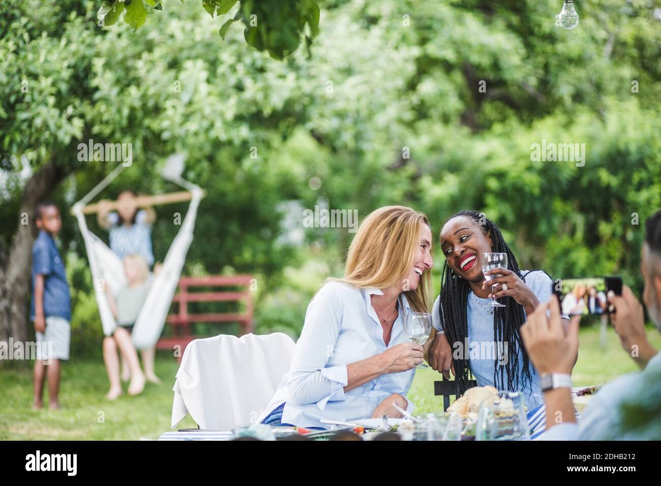 Uomo con telefono cellulare fotografando donne sorridenti che hanno vino dentro festa in giardino nel fine settimana Foto Stock