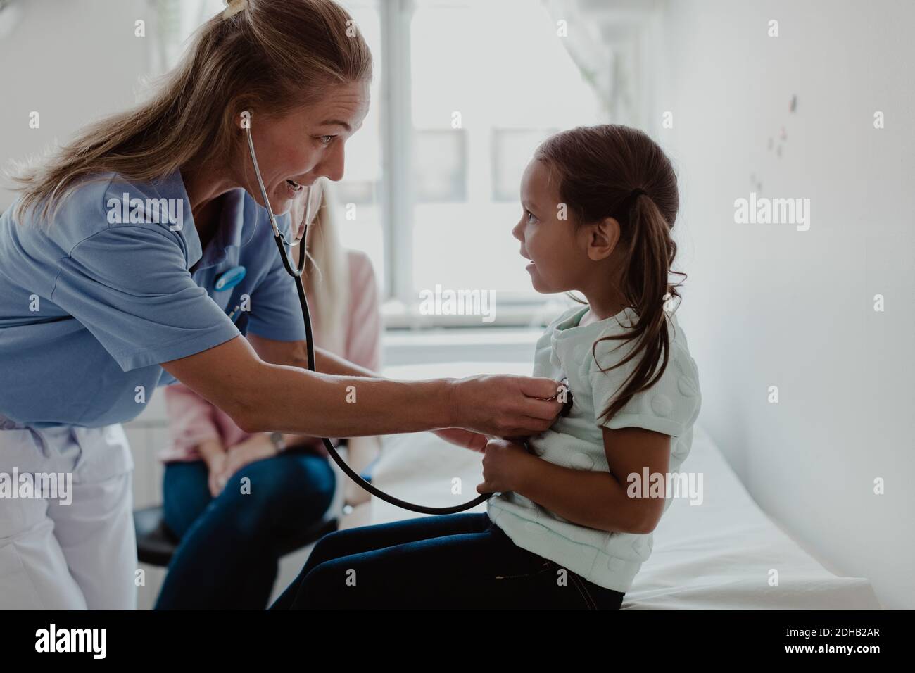 Pediatra femmina che esamina il battito cardiaco della ragazza attraverso stetoscopio mentre madre seduta in background in clinica Foto Stock