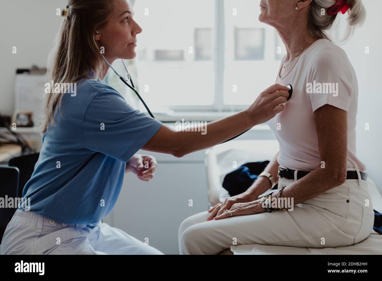 Vista laterale del medico femminile che ascolta il battito cardiaco della donna anziana in sala di visita medica Foto Stock