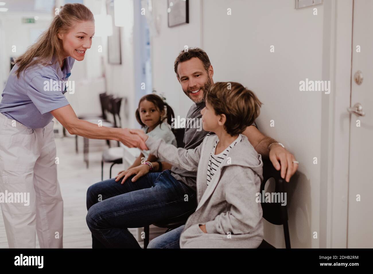 Sorridente pediatra femmina che agita con ragazzo seduto con la famiglia dentro corridoio ospedaliero Foto Stock
