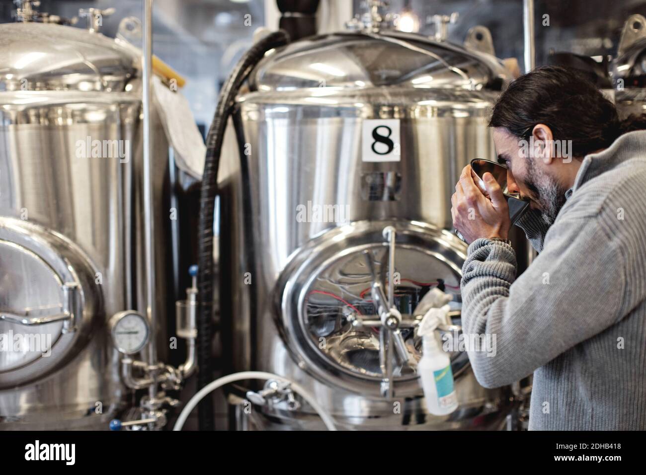 Uomo maturo che assaggia dal contenitore mentre si trova accanto al serbatoio di stoccaggio in birreria Foto Stock