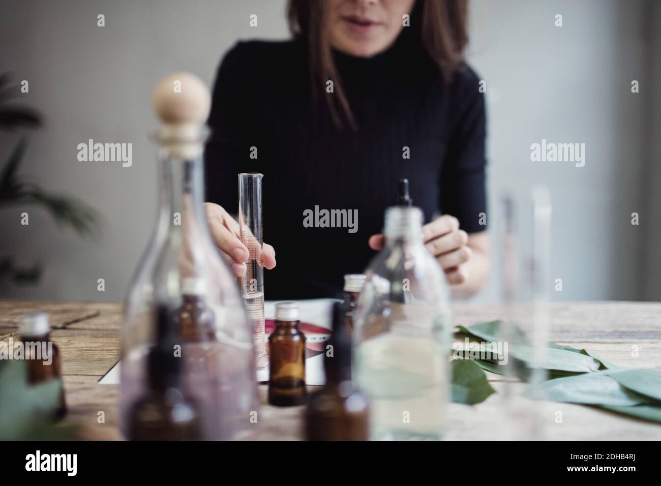 Sezione centrale del proprietario femminile che mescola il liquido mentre si siede al tavolo contro parete in laboratorio di profumo Foto Stock