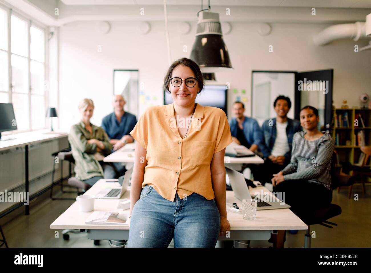 Ritratto di sorridente professionista appoggiato al tavolo contro dirigenti in un ufficio moderno Foto Stock