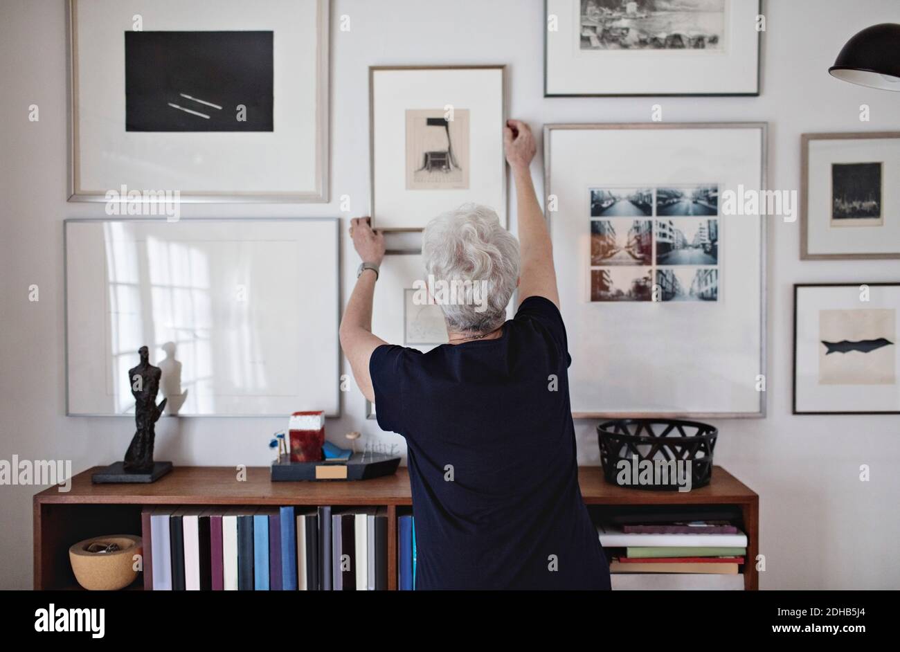 Vista posteriore della donna anziana ritirata che regola la cornice su parete sopra la libreria a casa Foto Stock