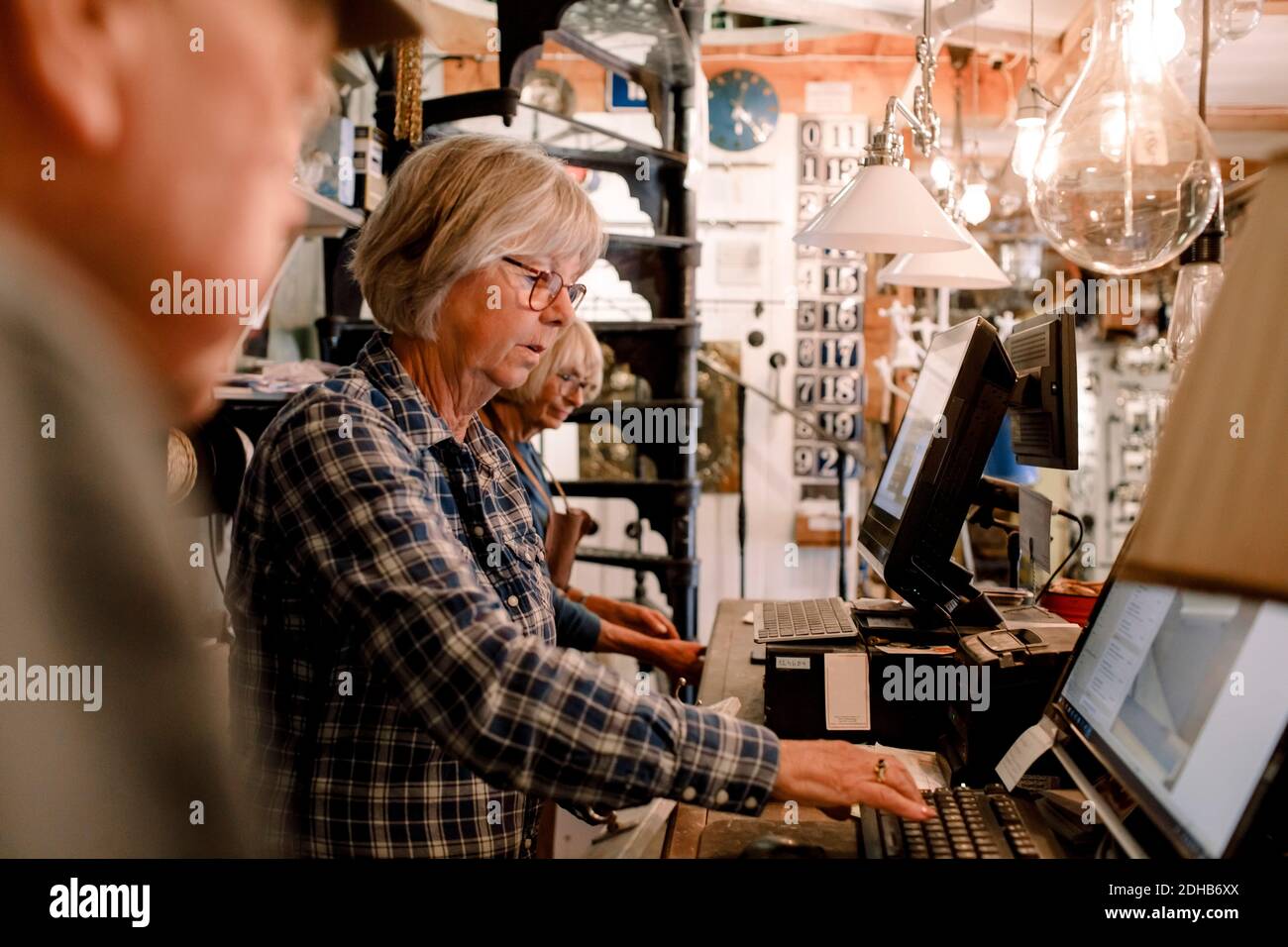 Donna senior sicura che usa il computer mentre si trova in mezzo a colleghi al momento del check-out in un negozio di hardware illuminato Foto Stock