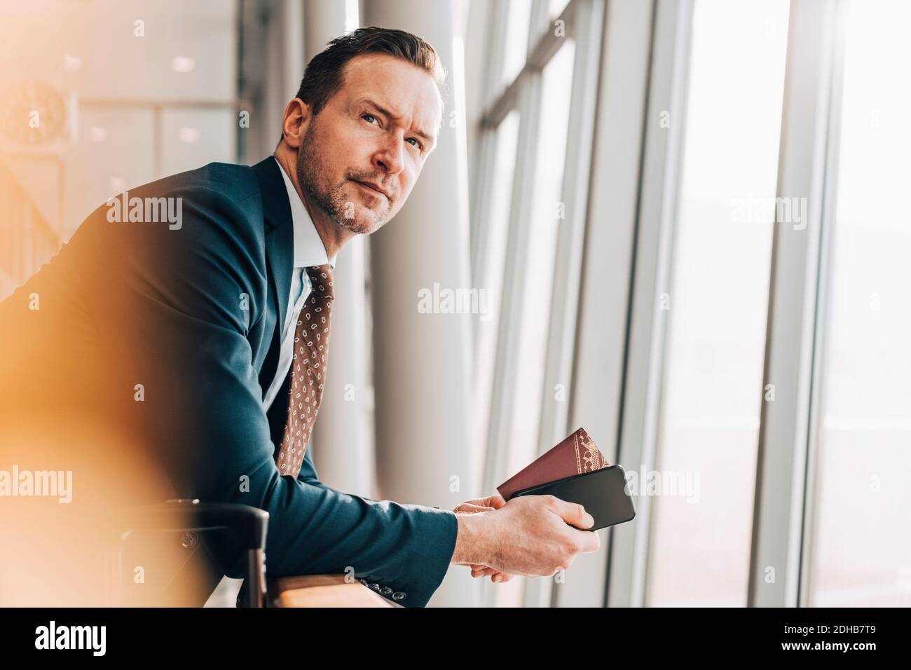 Uomo d'affari sicuro e maturo con passaporto e telefono cellulare appoggiato ringhiera all'aeroporto Foto Stock