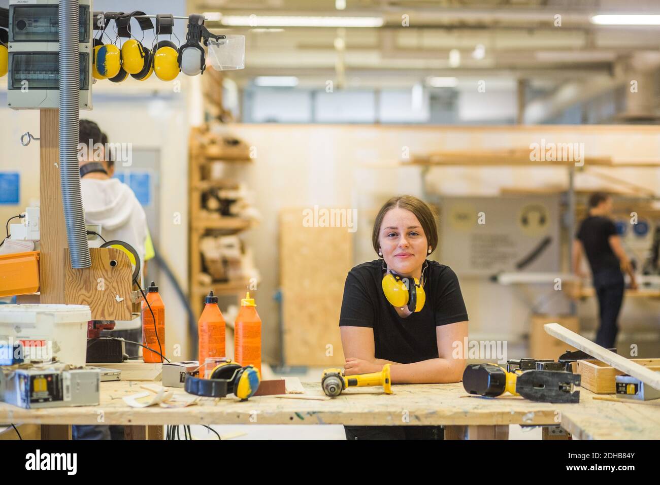 Ritratto sorridente giovane donna carpenteria tirocinante appoggiata su banco da lavoro con utensili elettrici in officina illuminata Foto Stock