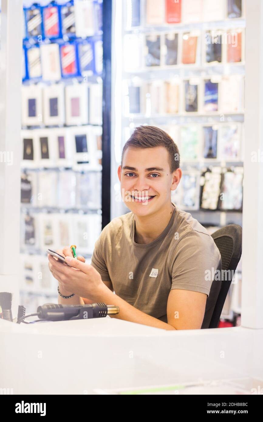 Ritratto di sorridente giovane uomo tirocinante riparazione telefono cellulare mentre seduto alla scrivania illuminata nel negozio Foto Stock