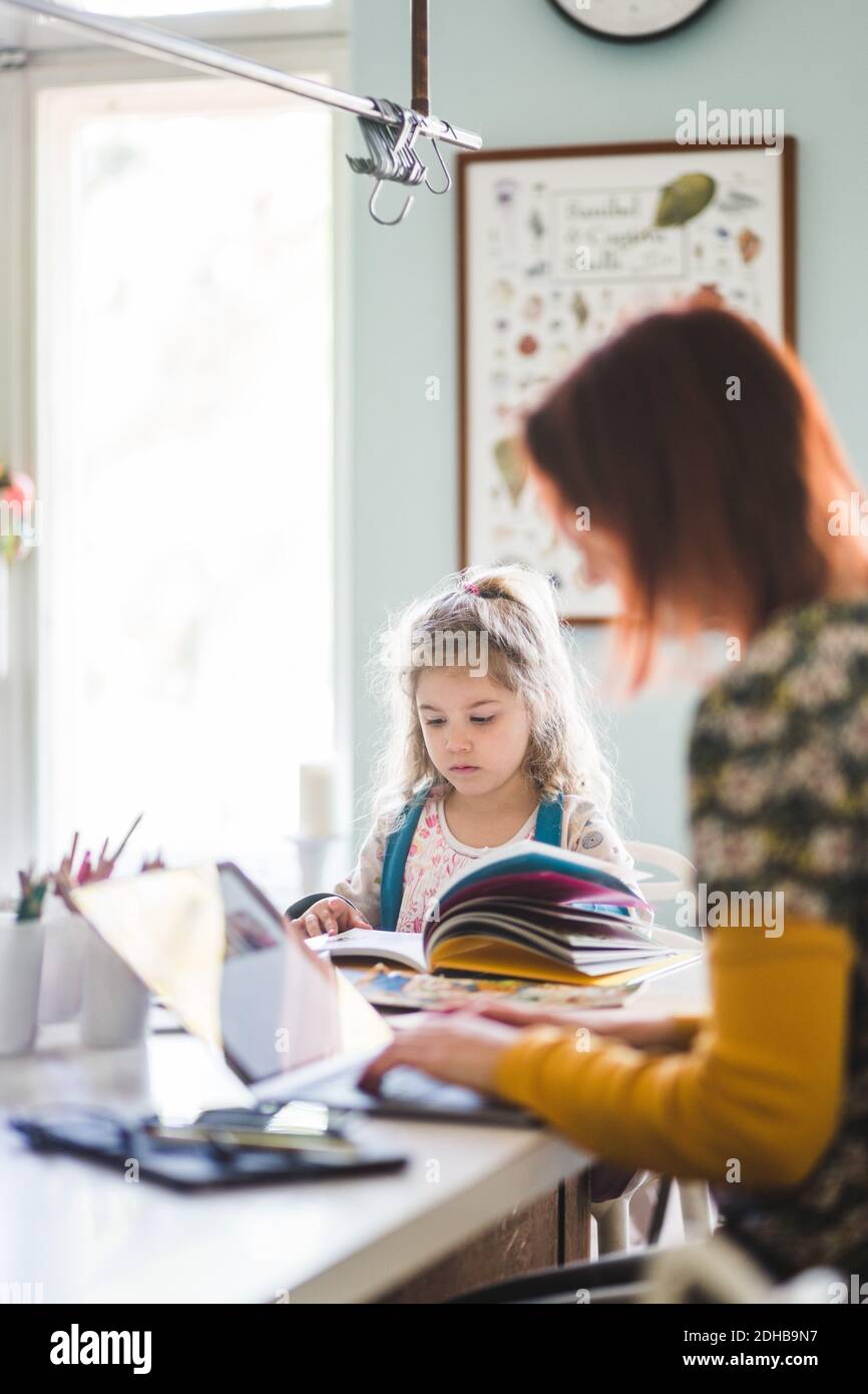 Vista laterale di una donna che usa un computer portatile mentre è seduto da una ragazza studiare all'isola della cucina Foto Stock