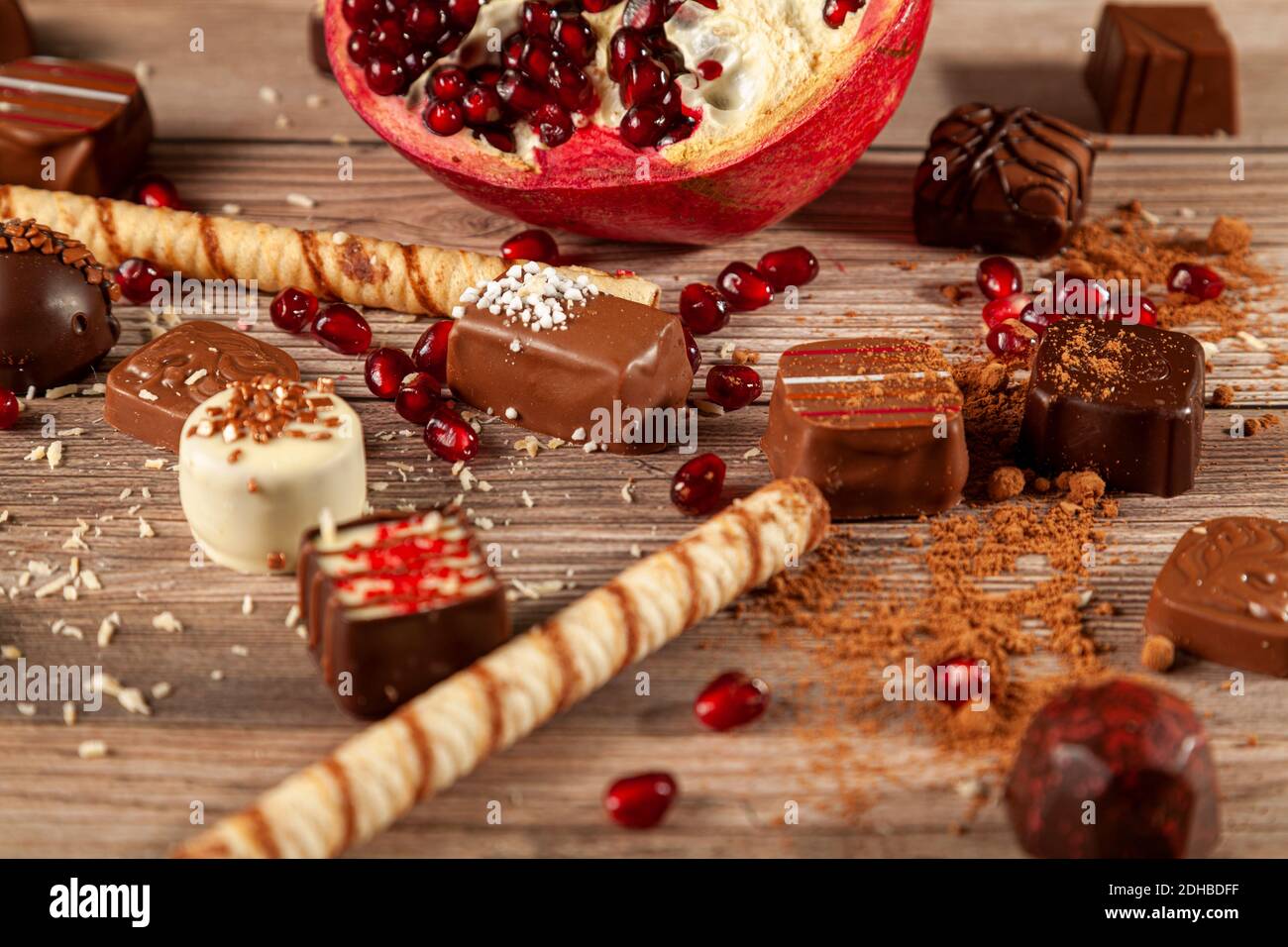 Semi di melograno affettato con semi sparsi su tavola di legno ricoperta di biscottini, cioccolatini assortiti, cacao in polvere e trucioli di cocco generi Foto Stock