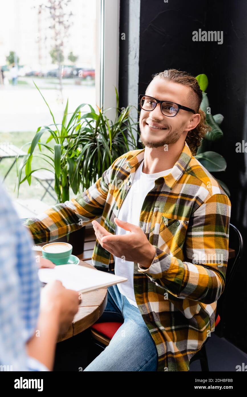 sorridente giovane uomo in occhiali puntando con un dito vicino al cameriere in primo piano sfocato Foto Stock