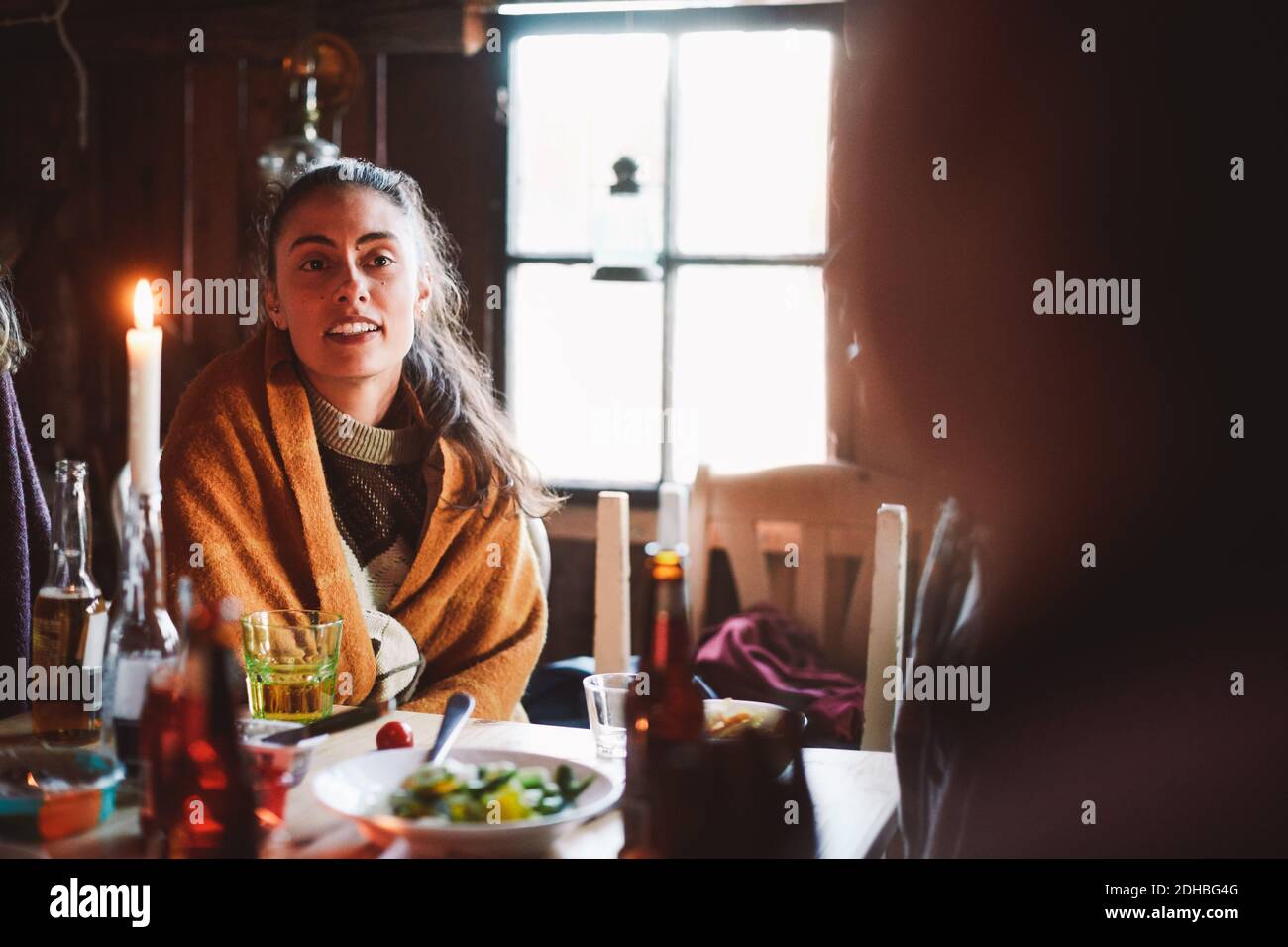 Giovane donna che indossa una coperta che guarda via mentre si siede al tavolo contro finestra in cabina di tronchi Foto Stock