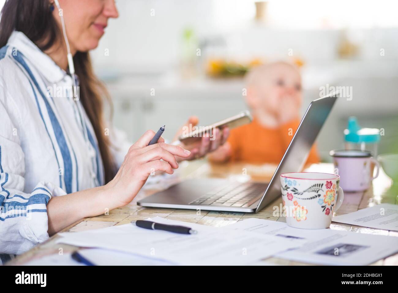 Sezione centrale della madre lavoratrice che utilizza tecnologie mentre la figlia si siede dentro sfondo al tavolo da pranzo Foto Stock