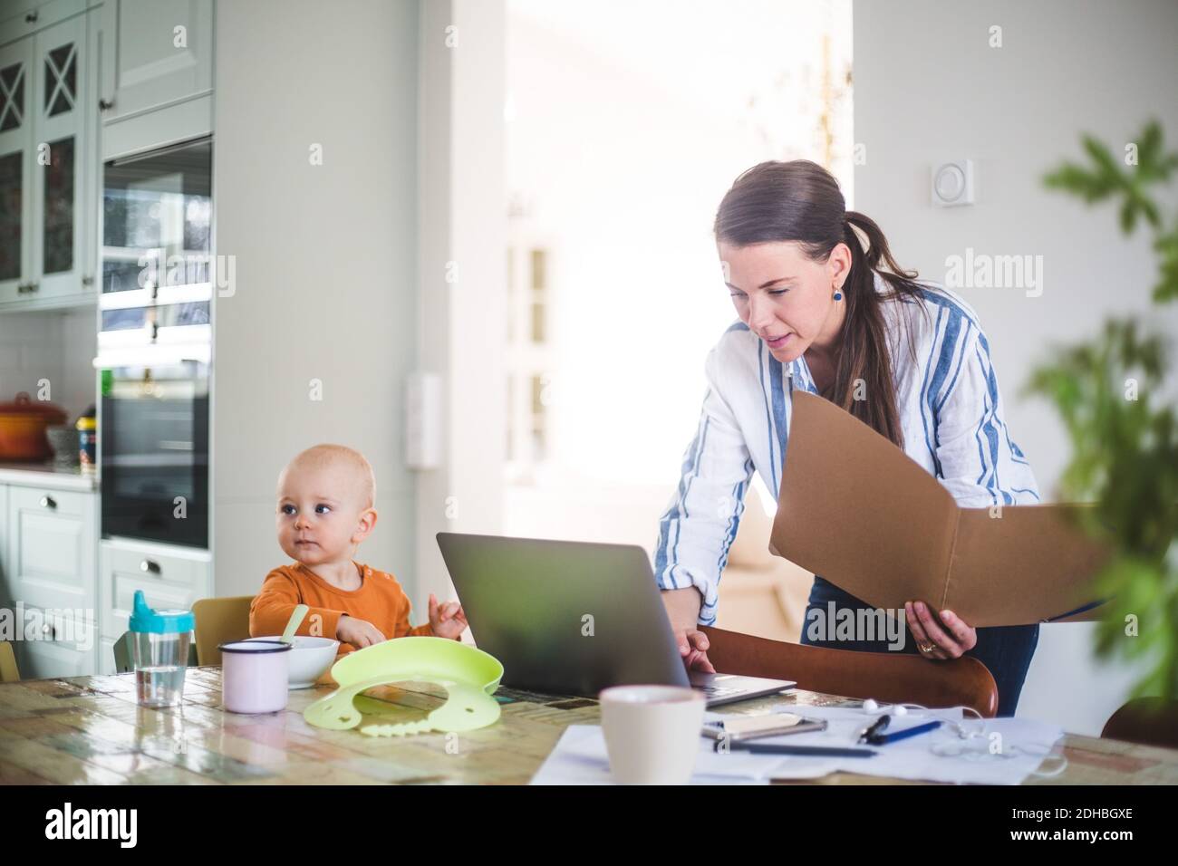 Femmina professionista che tiene file mentre si utilizza il computer portatile da figlia su tavolo da pranzo in ufficio a casa Foto Stock