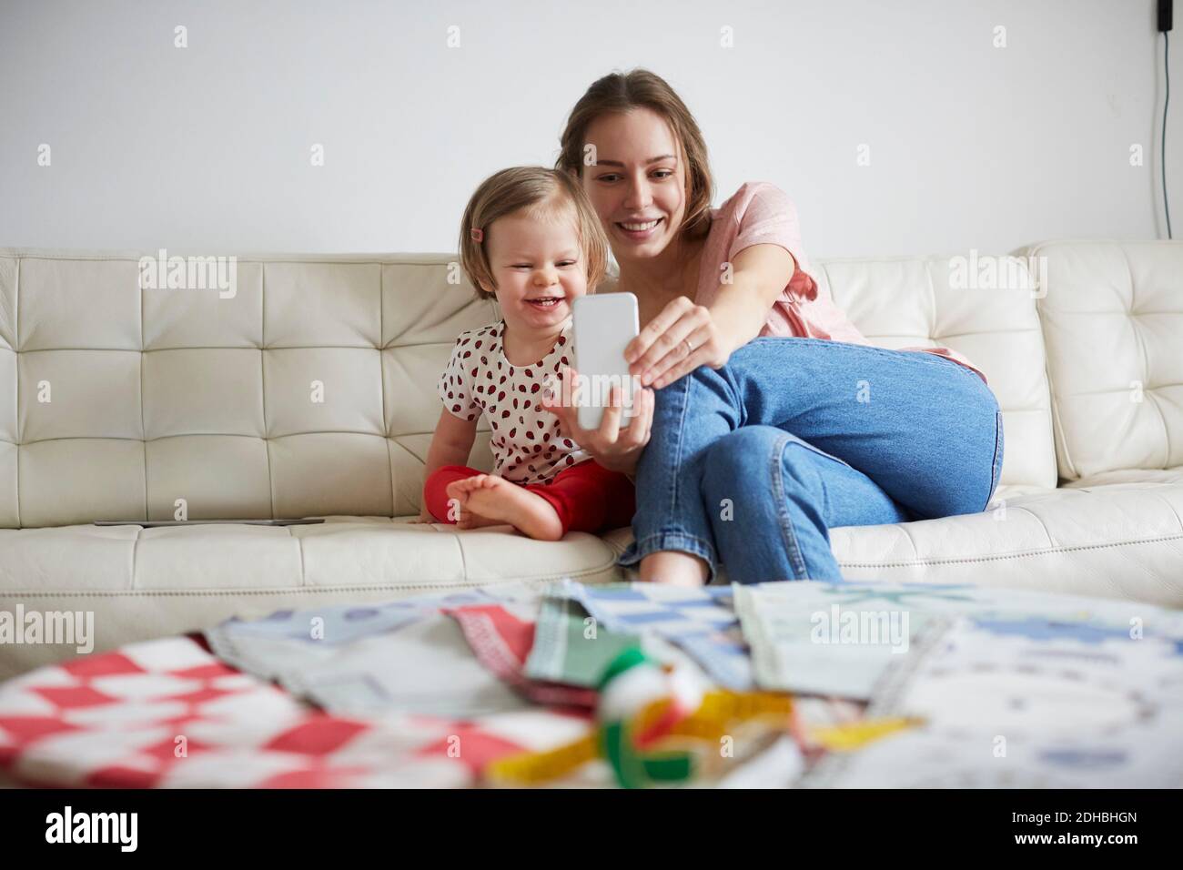 Sorridente madre e figlia che prendono selfie attraverso smartphone mentre seduto sul divano a casa Foto Stock