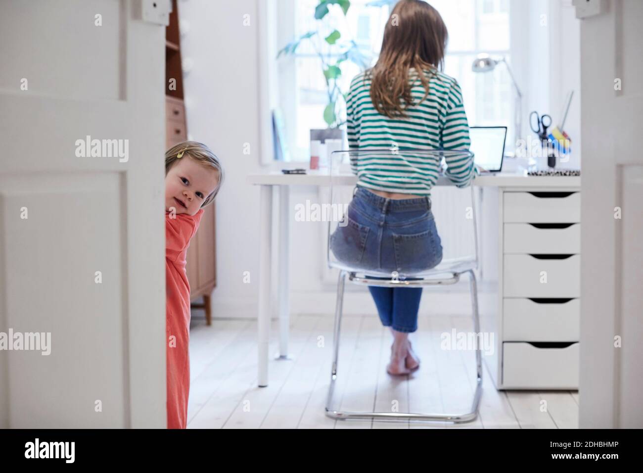 Ragazza in piedi dietro la porta mentre la madre lavora sul computer portatile sfondo a casa Foto Stock