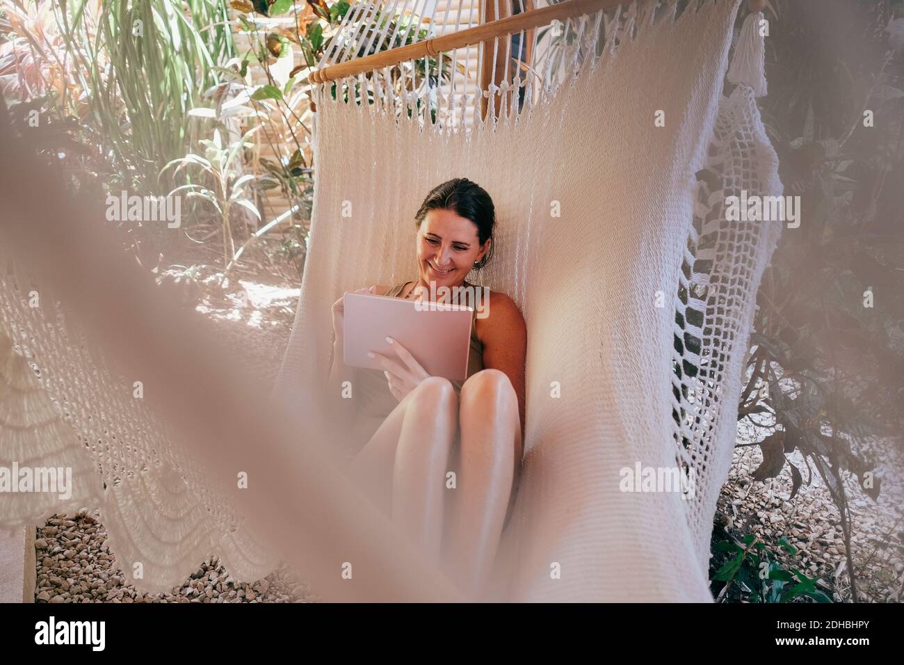 Donna matura sorridente che usa un tablet digitale in amaca sulla spiaggia Foto Stock