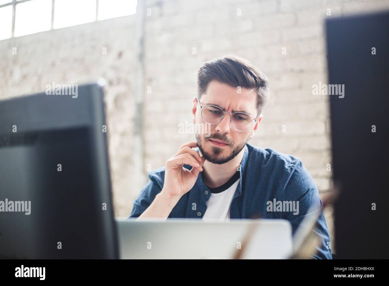 La barba è toccata da un giovane esperto IT mentre si guarda il computer portatile in ufficio Foto Stock